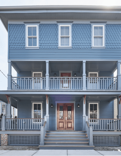 A large blue two-story house with a covered porch, white trim, and two front doors. The house features multiple windows and a fenced balcony on the second floor.