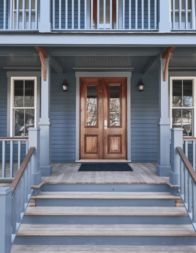 A blue-painted house with a wooden front porch, featuring double doors flanked by two windows. The porch has stairs with wooden railings leading up to the entrance.