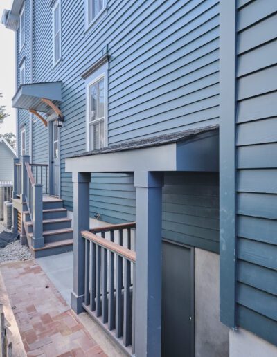 Side view of a blue wooden house featuring a small covered entryway to a basement, a side staircase, and a narrow brick path. The setting appears to be a residential backyard area.