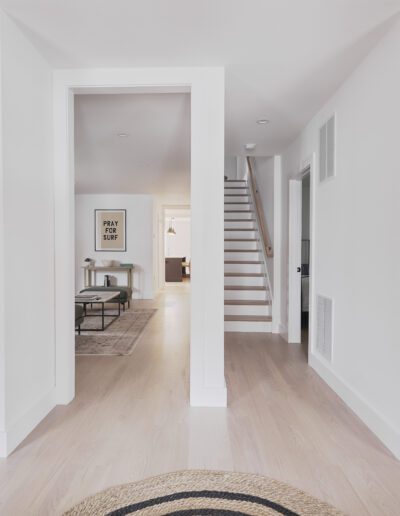 A modern, well-lit hallway with white walls and light wood flooring, featuring a staircase on the right and a living area visible through a doorway ahead. Artwork and rugs accent the space.