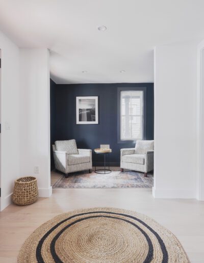 A modern entryway with wooden doors, a round woven rug, and a cozy seating area featuring two armchairs, a small table, and a framed picture on a dark accent wall.