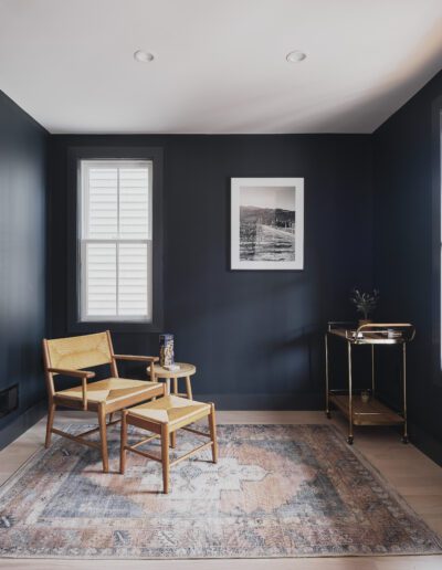 A small room with dark walls features a wooden chair and table on a patterned rug, a framed photograph on the wall, a window with white shades, and a bar cart in the corner.