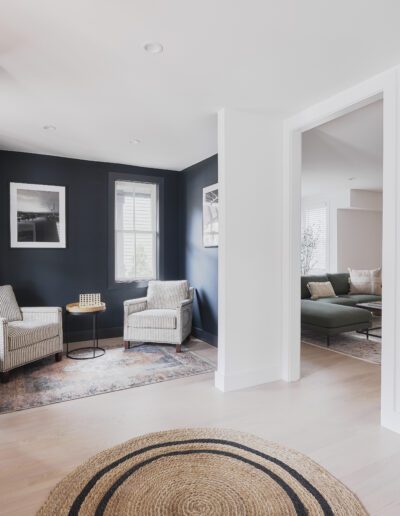 A modern, open living space with light wood flooring, featuring two beige armchairs and a small table against a dark accent wall. A circular woven rug and a wall mirror are in the foreground.