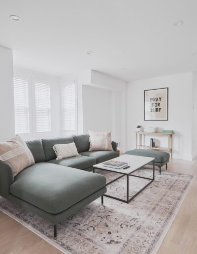 A modern living room with a green sectional sofa, a potted plant, a wooden side table, and a framed print on the wall. Light hardwood floors and large windows provide natural light. A kitchen is visible in the background.