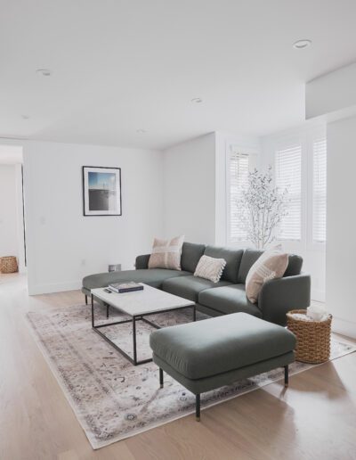 A modern living room with a green sectional sofa, a coffee table on a patterned rug, white walls, and large windows with white blinds. A hallway leads to a door in the background.