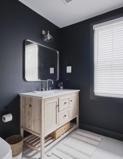 A modern bathroom with dark blue walls, a wooden vanity with a marble countertop, a rectangular mirror, a white toilet, and a window with white blinds.