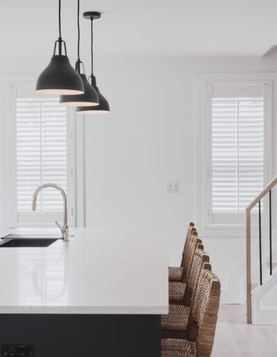 A modern kitchen with white walls and countertops, a central island with wicker bar stools, three black pendant lights, and a staircase to the right. White shutters cover the windows.