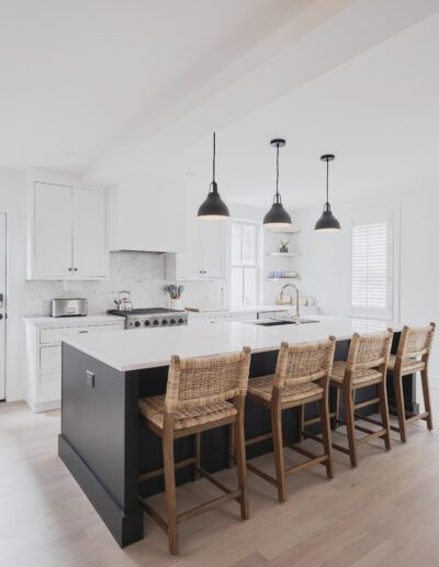 A modern kitchen with a large island, wicker bar stools, hanging pendant lights, white cabinets, stainless steel appliances, and a door leading outside.