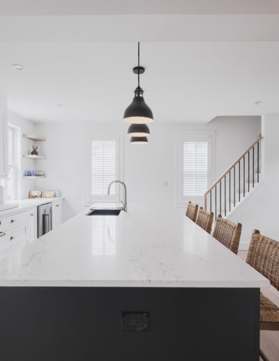 Modern kitchen with white cabinets, a large marble island, stainless steel appliances, and wicker barstools. Hanging lights are above the island, and a dining table is visible in the background.