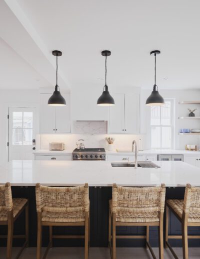 A modern kitchen featuring a large island with a white countertop, four wicker chairs, and three black pendant lights. Stainless steel appliances are visible, and the space is brightly lit by natural light.