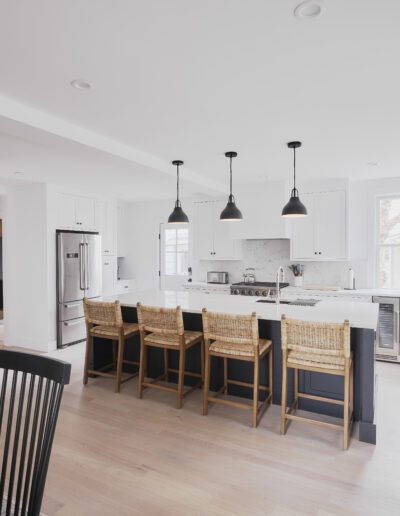 A modern kitchen with white cabinets, a large island with wooden barstools, stainless steel appliances, and three pendant lights. Adjacent to it is a dining area with a table and black chairs.