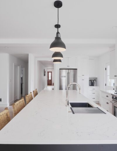 Modern kitchen with a long white island, wicker barstools, pendant lights, stainless steel appliances, and white cabinetry. An adjacent dining area with a table and black chairs is visible in the background.