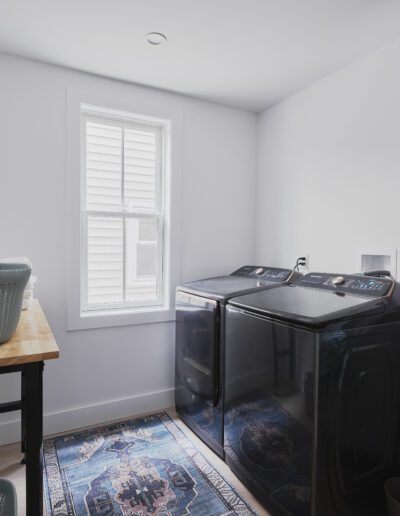 A laundry room with a washer and dryer, a wooden table with a basket, an ironing board, and shelves with folded towels. A window lets in natural light, and the floor has a patterned rug.