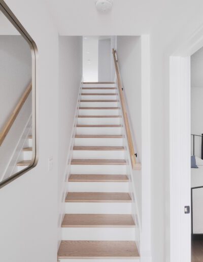 A bright, modern hallway with wooden stairs leading up. A mirror is mounted on the left wall. A bedroom with a black metal bed frame is visible through an open door on the right.