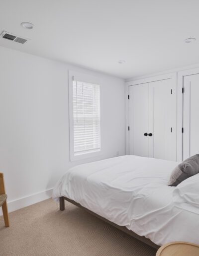 A minimalist bedroom with a white bed, wooden chair, small round side table, and built-in white closets, featuring a window with blinds.