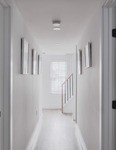 A hallway with white walls, wooden flooring, and framed pictures on the walls. Doors on either side lead to other rooms, and a window at the end lets in natural light. A staircase is visible at the far end.