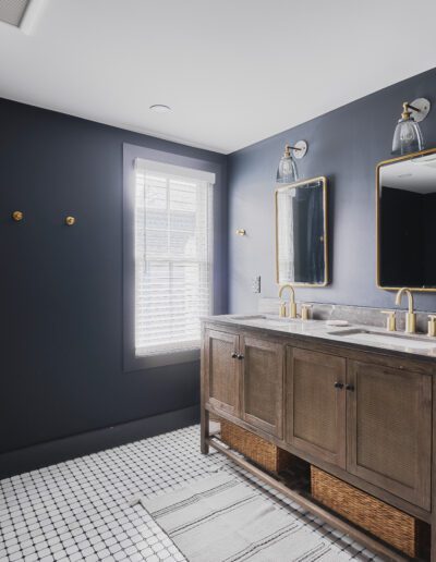 A modern bathroom features a double sink vanity with gold fixtures, two mirrors, dark walls, a white tiled floor, a window with blinds, and a towel rack on the wall.