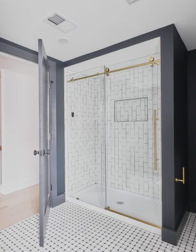 A modern bathroom with black walls featuring a glass shower enclosure with gold accents and a white toilet. The floor is tiled in black and white. The door is partially open, showing a hallway.