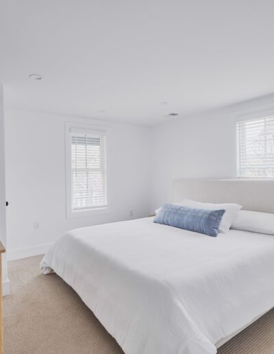 A minimalist bedroom with white walls, a large bed with white bedding and a blue accent pillow, a wooden dresser with a remote control on it, and two windows with white blinds.