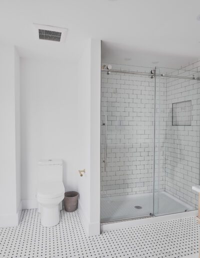 A modern bathroom features a glass-enclosed shower with white subway tiles, a white toilet, a wooden bench, a vanity with a marble countertop, and a window with blinds.