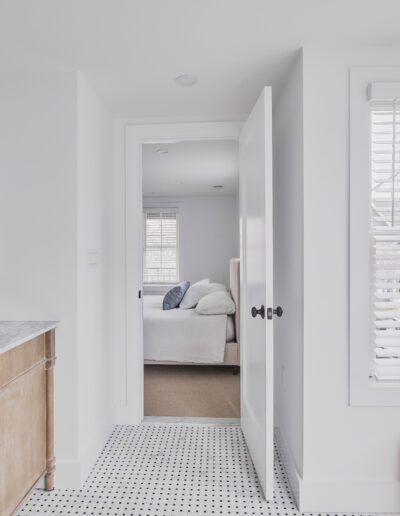 Minimalist bathroom with a marble countertop, wooden vanity, and brass fixtures. A door leads to a bedroom with a made bed. A wicker basket and a window with blinds are on the right.