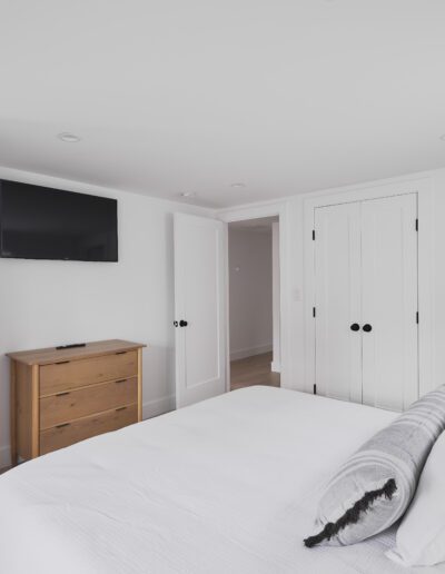 Minimalist bedroom with white walls, a mounted flat-screen TV, wooden dresser, double bed with white and gray bedding, two closets, and an open door leading to a bathroom.