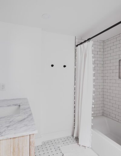 A modern bathroom with white tiled bathtub, black shower rod and curtain, marble countertop with sink, black faucet, wall mirror, and light fixture.