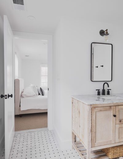 Minimalist bathroom with a wooden vanity, marble countertop, a black faucet, and a rectangular mirror. Open door reveals a glimpse of a bedroom with a white bed.