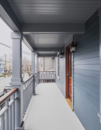 A covered porch with blue siding, a wooden front door, and white flooring overlooks a residential street lined with parked cars and trees.