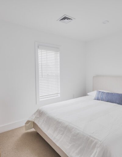 A minimalist bedroom with white walls, a double bed with a white duvet and blue pillow, a wooden dresser, a window with blinds, beige carpet, and a ceiling vent.