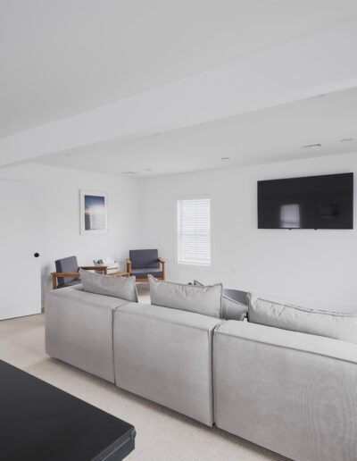 Modern living room with a light gray sectional sofa facing a wall-mounted flat-screen TV, two armchairs and a small table in the background, and bright white walls with windows.