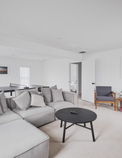 A well-lit living room featuring a gray sectional sofa, a round black coffee table, two gray armchairs, a dining table in the background, and two framed pictures on white walls.