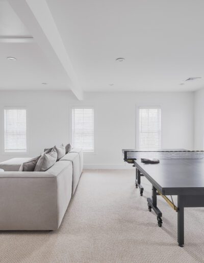 A spacious room with a gray sofa, black coffee table, wall-mounted TV, and a ping pong table. The room has white walls, beige carpet, and three large windows with blinds. A framed picture hangs on the wall.