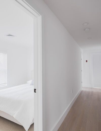 Hallway with wooden floor leading to a bright bedroom with white walls and bed. A staircase with a black handrail is on the right side. The bedroom door is open, revealing a window with closed blinds.