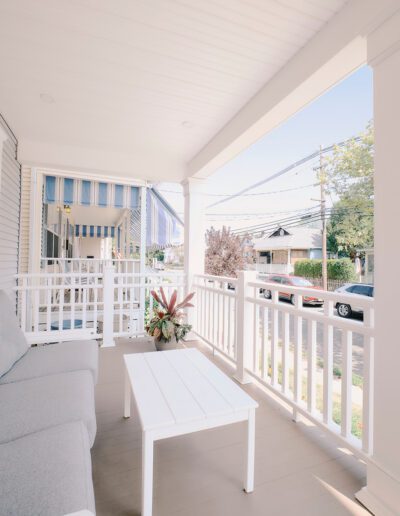 A view of a bright porch with a white railing, a white coffee table, a potted plant, and a grey cushioned bench. The scene includes a glimpse of neighboring houses and the street in the background.