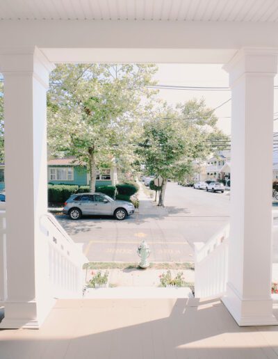 View from a covered front porch showing white stairs leading down to a sidewalk, a parked car, trees, and houses across a sunny street.