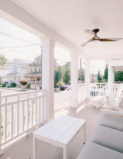 A white porch with a ceiling fan, cushioned seating, and tables overlooks a suburban street with trees and homes.