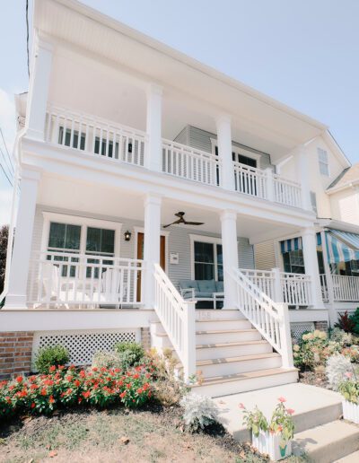 Two-story house with a spacious front porch and balcony, white railings, and a well-maintained front garden featuring various flowering plants. The house number "138" is visible on the steps.