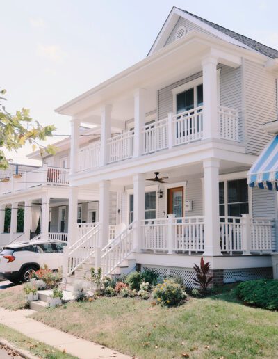 A two-story house with a white exterior, two front porches, and a manicured lawn. A white car is parked in the paved driveway to the left of the house.