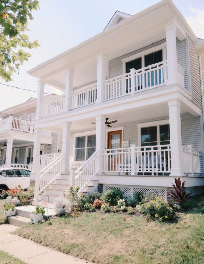 A two-story house with a double porch, white railings, light gray siding, and a small garden in the front yard.