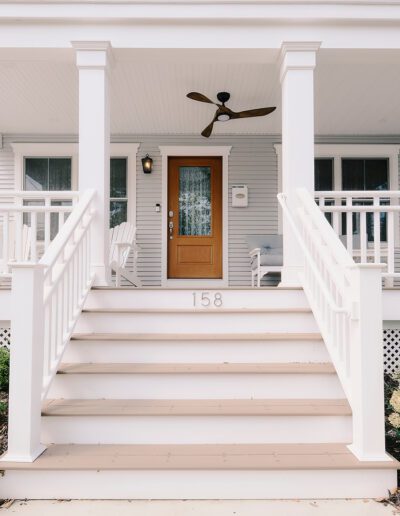 Front view of a house with a wooden door, white railing, stairs, and a porch with a ceiling fan. The house has light blue siding and is adorned with plants and flowers in the front garden.