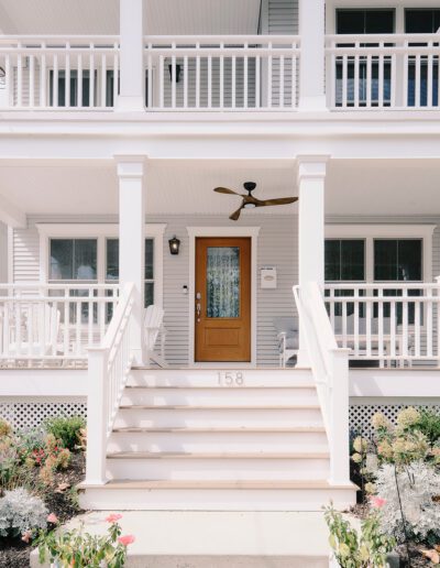 Front view of a two-story house featuring a wooden front door, white columns, and a porch with railings. Steps lead up to the entrance with potted flowers on each side and the number 158 on the step.