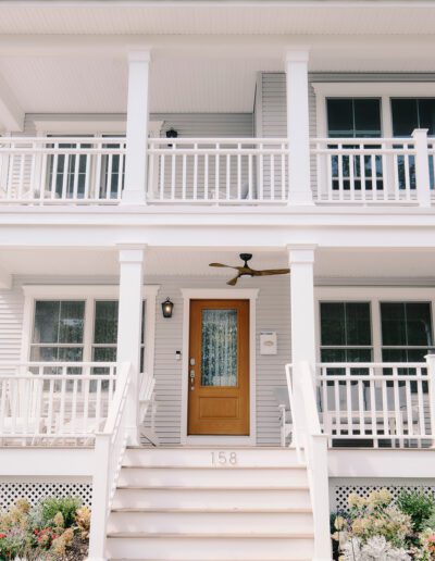 Two-story white house with a wooden front door, front porch, and upper balcony. The house number "158" is displayed on the front steps. The front yard has flowers and greenery.