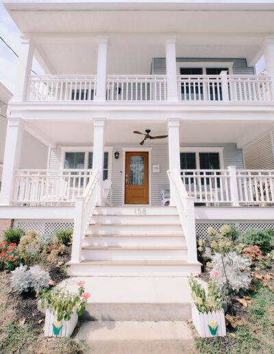 A two-story white house with a large front porch, detailed railings, a wooden front door, ceiling fan, and outdoor landscaping including flowers on either side of the entrance steps.