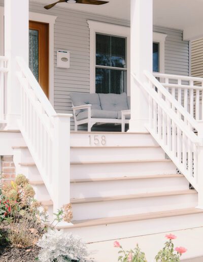 White front porch with a wooden door, light fixture, and a set of stairs with railing leading to it. The house number 158 is on the stairs. Flowered garden beds line both sides.