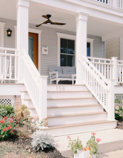 Front porch of a light grey house with white railings, stairs, ceiling fan, and address number "158." There are potted flowers and a small garden in front. Two cushioned chairs sit on the porch.