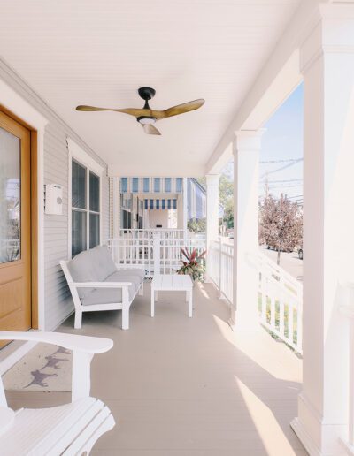 A well-lit, white wooden porch with Adirondack chairs, a small table, and a ceiling fan overlooking a street with parked cars and trees.