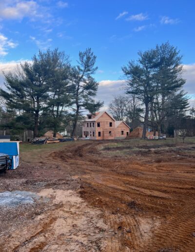 A house under construction surrounded by trees and dirt, with scattered building materials and a clear blue sky in the background.