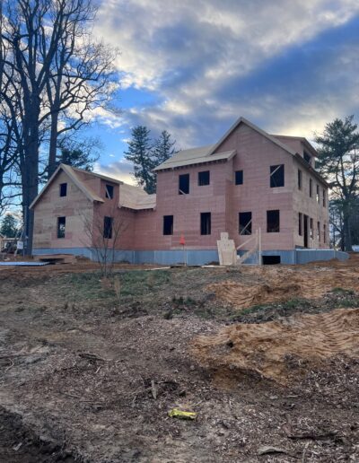 A two-story house under construction, with the exterior framework and roof partially built, situated on a cleared plot of land with trees in the background.