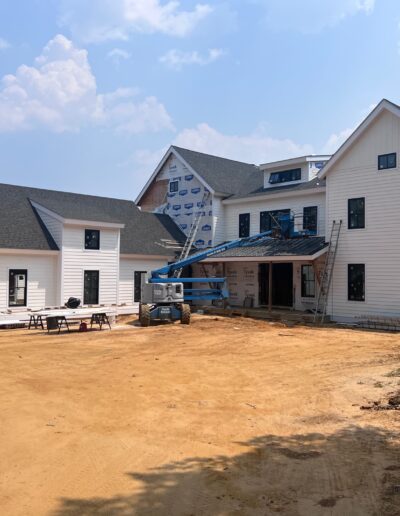 A large house under construction with white exterior walls and black roofs. A lift and construction materials are present in the driveway. A red car is parked on the left side.